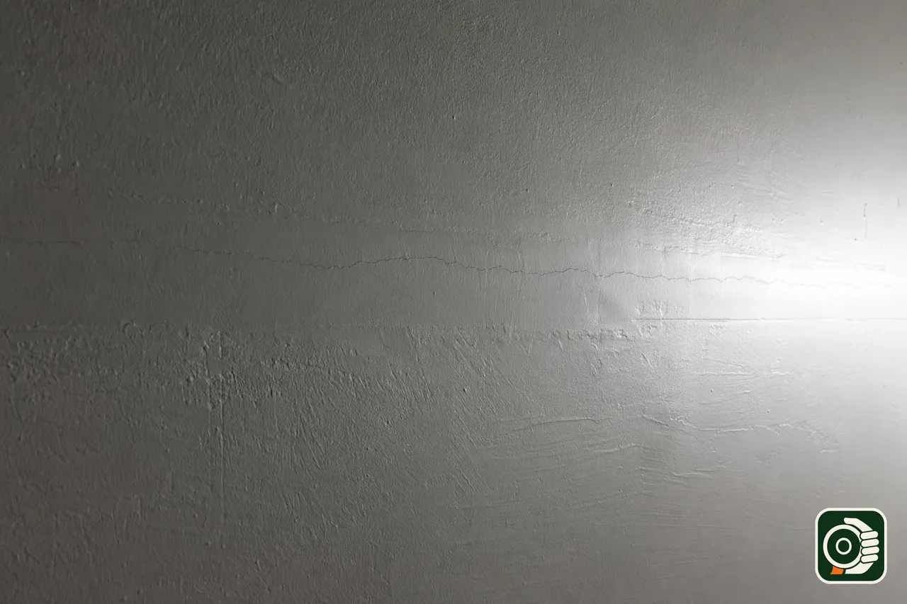 Parete in cartongesso illuminata di taglio: la luce radente rivela una microcrepa lungo il giunto e le righe della spatola nella rasatura